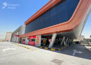 Property for sale A modern commercial building with a curved red facade, large glass windows, and angled pillars. At ground level are fast-food restaurants and parking spaces, offering views reminiscent of DAMAC Riverside Views. Clear directional arrows guide visitors. Presented by Palace Auctions