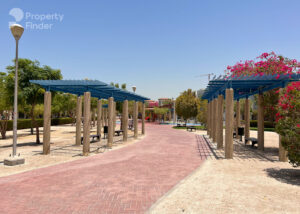 Property for sale A paved walkway curves through a sunny park with wooden pergolas, benches, trees, and flowering shrubs—perfect for residents of a luxury flat in Jumeirah Village Triangle, Dubai. Lamp posts and a children’s play area complete the inviting scene. Presented by Palace Auctions