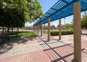 Property for sale A walkway with tall beige columns and a blue pergola casting shadows, surrounded by trees and bushes in Jumeirah Village Triangle, Dubai. Tennis courts are visible in the background near a luxury flat on a sunny day. Presented by Palace Auctions