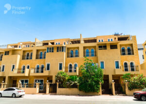 Property for sale A row of yellow, multi-storey townhouses with balconies and arched windows, lush greenery in front, and parked cars on the street under a clear blue sky—an inviting setting for a luxury JVC Smart Apartment or Dubai investment flat 2026. Presented by Palace Auctions