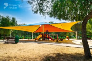 Property for sale A sunny playground with colourful slides and climbing equipment is shaded by large yellow canopies, surrounded by trees in a green park—perfect for families living in nearby Luxury JVC Smart Apartments or seeking a Dubai smart home property. Presented by Palace Auctions