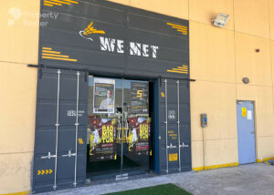 Property for sale A shopfront with a black and grey entrance labelled "WE NET," decorated with badminton posters and yellow accents, stands against a beige wall near DAMAC Riverside Views, featuring a blue utility door and a CCTV camera above. Presented by Palace Auctions