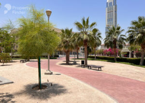 Property for sale A sunny park with palm trees, a winding red-brick path, benches, and a tall modern skyscraper in the background evokes Dubai’s vibrant atmosphere near Jumeirah Village Triangle under a clear blue sky. Presented by Palace Auctions