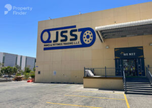 Property for sale A beige industrial building near Capri 2 with a large blue and yellow "JSS Pipes & Fittings Trading LLC" sign above the loading bay and entrance on a sunny day. The Property Finder watermark is in the top left corner. Presented by Palace Auctions