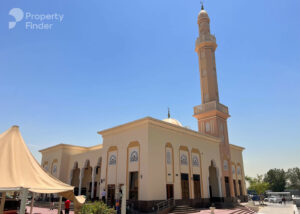 Property for sale A beige mosque with decorative arches, tall minaret, and dome stands under a clear blue sky near DAMAC Riverside. Several cars are parked nearby, a tent canopy is to the left, and the Property Finder logo appears in the top left corner. Presented by Palace Auctions