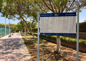 Property for sale A shaded park path with benches and trees, near a luxury block of flats in Jumeirah Village Triangle, Dubai. A large sign in English and Arabic lists community rules, whilst a person walks by a green fence in the distance. Presented by Palace Auctions