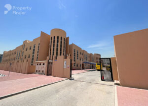 Property for sale A modern beige building, Capri 2, with tall rounded towers and many windows stands by a pink-tiled walkway and black metal gate. Inside, a yellow coach is parked near a blue canopy, offering serene DAMAC Riverside Views under a clear blue sky. Presented by Palace Auctions