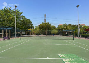 Property for sale A clean, empty outdoor tennis court in Jumeirah Village Triangle, Dubai, surrounded by trees and fences under a clear blue sky, with a few benches and tall light poles visible near luxury flat buildings in the background. Presented by Palace Auctions