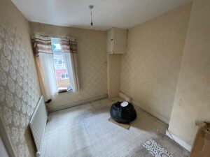 Property for sale A small, empty room with patterned wallpaper, a window with curtains, a radiator, and a worn carpet. The Stanley property for sale features a ceiling light fitting without a shade, a black bag on the floor, and a cupboard in the corner. Presented by Palace Auctions