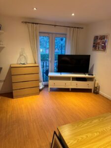 Property for sale A tidy living room in a prestigious development in Weybridge, featuring wooden flooring, a white TV stand with a flat-screen telly, light wood chest of drawers, white curtains over glass doors, and shelves with decor on the left wall in this 2-bed flat. Presented by Palace Auctions