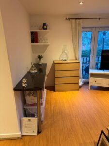 Property for sale A tidy room in a prestigious development in Weybridge, featuring wooden floors, black worktop with shelves above, kettle and mugs, light wood chest of drawers, large vase, and glass doors with white curtains on the right. Presented by Palace Auctions