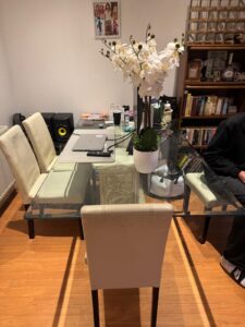 Property for sale A glass dining table with cream chairs, a white orchid centrepiece, a laptop, books, and speakers sits in this 2-bed flat in Weybridge’s prestigious development. Bookshelves and framed photos are in the background with part of a person seated on the right. Presented by Palace Auctions