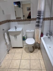 Property for sale A bathroom in a 2-bed flat within a prestigious development in Weybridge, featuring a washbasin with cupboard, wall mirror, loo, towel on the left, cleaning products beside the loo, and a bath with striped shower curtain. Tiled flooring throughout. Presented by Palace Auctions