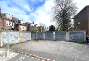 Property for sale A row of three grey garage doors with graffiti stands beside a paved area, bordered by brick buildings, a fence, and a large tree under a partly cloudy sky—ideal for planning or development site opportunities in Winchmore Hill N21. Presented by Palace Auctions