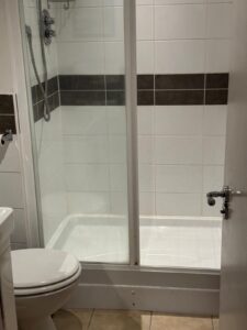 Property for sale A small bathroom in a prestigious development in Weybridge, featuring a glass shower enclosure, white tiles with a dark stripe, a silver showerhead, closed white toilet, and a partially open white door—ideal for this modern 2-bed flat. Presented by Palace Auctions