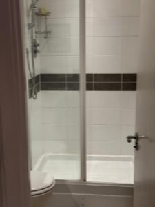 Property for sale A bathroom in a prestigious development in Weybridge, featuring a glass-enclosed shower with white tiles and a horizontal dark stripe, a modern showerhead, and part of the 2-bed flat’s loo visible in the foreground. Presented by Palace Auctions