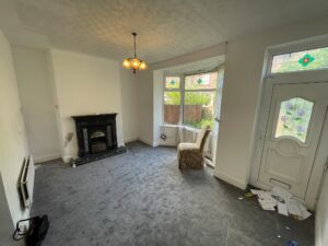 Property for sale A sparsely furnished living room with grey carpet, a fireplace, and a single armchair by a bay window in this Stanley property for sale. Some papers and rubbish are scattered near the white front door; light fitting hangs from the ceiling. Presented by Palace Auctions