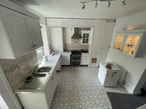 Property for sale A small kitchen in a Stanley 3-bed terrace with white cupboards, patterned tile flooring, a double sink, gas cooker with oven and hood, fridge, and a window for natural light. Minimalist worktops and white walls complete this Stanley property for sale. Presented by Palace Auctions