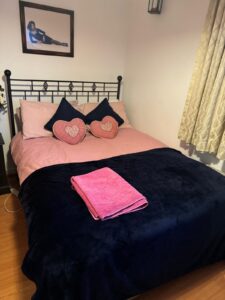 Property for sale A neatly made bed with a pink sheet, navy blue blanket, and two pink heart-shaped cushions sits against a wall in this prestigious development. Folded pink towels are on the bed, and a framed photo hangs above it in this stylish Weybridge two-bed flat. Presented by Palace Auctions