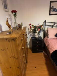 Property for sale A cosy 2-bed flat in a prestigious development, featuring a wooden chest of drawers, gold horse statue, and red roses. A black bedside table holds mixed flowers beside the bed with pink bedding and framed art, creating a stylish Weybridge retreat. Presented by Palace Auctions