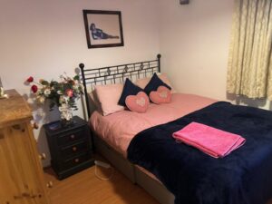 Property for sale A neatly made bed with pink sheets, navy blue blanket, and heart-shaped cushions sits in a cosy 2-bed flat in prestigious development, Weybridge. A vase of flowers and a framed photo decorate the bedside table; folded towels rest on the bed. Presented by Palace Auctions