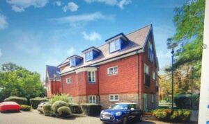 Property for sale A modern red-brick block of flats with dormer windows, set in greenery within a prestigious development in Weybridge. Several cars, including a blue Mini, are parked in front under a bright, partly cloudy sky. Presented by Palace Auctions
