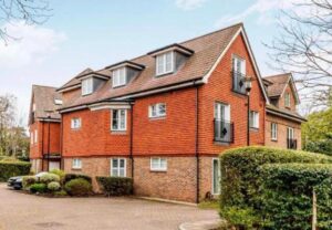 Property for sale A modern two-storey brick and red-tiled block of flats with dormer windows, set in a prestigious development in Weybridge, surrounded by hedges and greenery under a partly cloudy sky. Presented by Palace Auctions