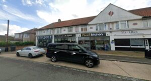 Property for sale A row of shops on a street, including a barber, a bar, a kitchen restaurant, and a hairdresser. Several parked cars are in front, and two people sit outside the restaurant under a cloudy sky. Presented by Palace Auctions
