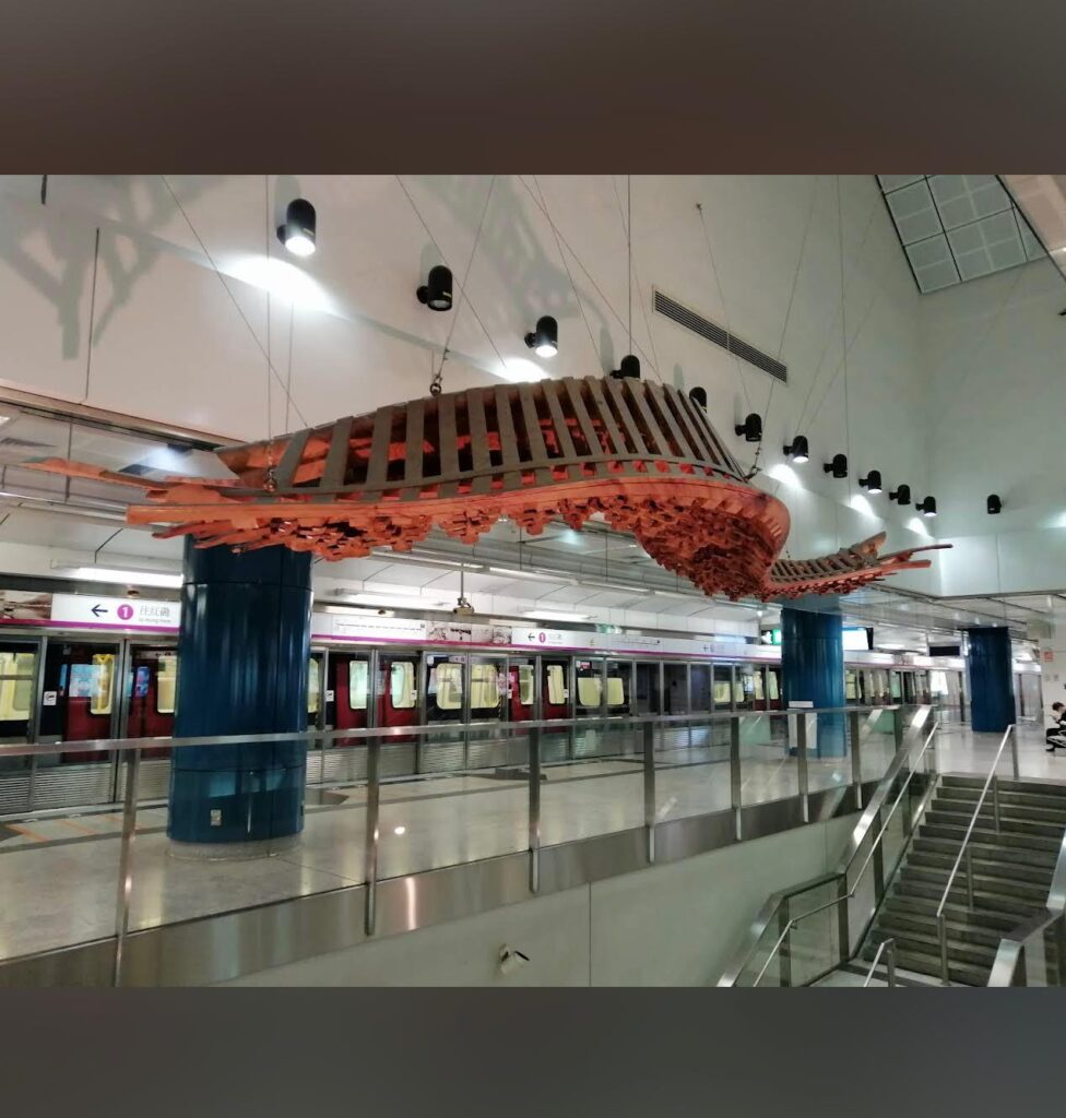 Property for sale A large wooden sculpture resembling a whale skeleton hangs from the ceiling of an underground station in Tuen Mun, above a platform with a red and white train and blue columns—perfectly blending urban art near your ideal Tuen Mun flat for sale. Presented by Palace Auctions