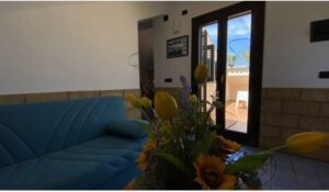 Property for sale A cosy living room with a blue sofa, a floral arrangement featuring yellow tulips and sunflowers, a door leading to a sunny patio, and a wall with tan tiles and a framed photo. Presented by Palace Auctions