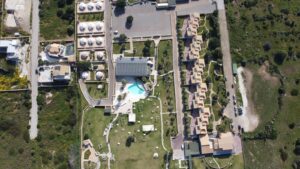 Property for sale Aerial view of a resort with a central pool, garden, and pathways, surrounded by multiple buildings and dome-shaped structures, set in a green landscape. Cars are parked near the entrance on the right-hand side. Presented by Palace Auctions