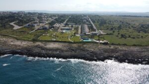 Property for sale Aerial view of a coastal resort with several buildings, green lawns, and swimming pools next to a rocky shoreline and the sea, with a rural landscape and scattered houses in the background. Presented by Palace Auctions