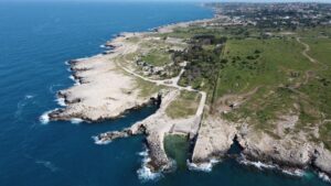 Property for sale Aerial view of a rocky coastline with clear blue sea, green fields, and scattered trees. A small inlet and stone pier extend into the water, with distant buildings and coastline visible on the horizon. Presented by Palace Auctions