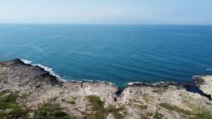 Property for sale Aerial view of a rocky coastline with gentle waves meeting the shore, clear blue sea stretching to the horizon under a clear sky. Presented by Palace Auctions