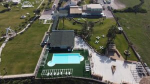 Property for sale Aerial view of a resort with a rectangular swimming pool, green lawns, scattered palm trees, outdoor seating areas, and several buildings, surrounded by open grassy fields. Presented by Palace Auctions
