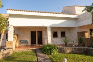 Property for sale A modern, single-storey house with stone walls, dark wooden doors, small rectangular windows, and a tiled roof. There are tropical plants and neatly trimmed grass in the front garden under bright sunlight. Presented by Palace Auctions