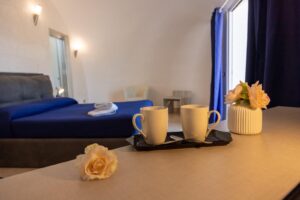 Property for sale A cosy bedroom with a neatly made bed covered in blue linen, two white mugs on a tray, and a vase with pink flowers on a table in the foreground. Sunlight enters through a window with blue curtains. Presented by Palace Auctions
