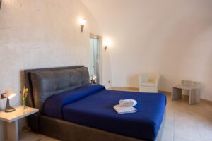 Property for sale A minimalist bedroom with a dark grey bed frame, navy blue bedding, folded towels on the bed, two bedside tables with lamps, a white armchair, and a small marble table against a textured white wall. Presented by Palace Auctions