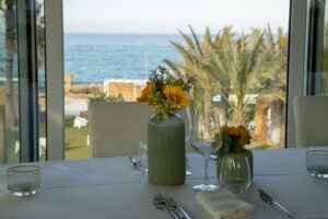 Property for sale A table laid for a meal with a white tablecloth, glassware, and green vases holding yellow flowers, positioned by large windows overlooking palm trees and the sea. Presented by Palace Auctions