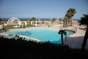 Property for sale A large outdoor swimming pool with clear blue water, surrounded by palm trees, white chairs, and decorative arches on a green lawn, overlooking the sea under a clear blue sky. Presented by Palace Auctions
