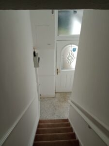 Property for sale View from the top of a staircase leading down to a white front door with a frosted glass panel and geometric pattern. The hallway has tiled flooring, a wall-mounted fuse box, and white walls. Presented by Palace Auctions