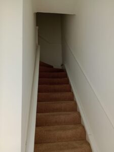Property for sale Carpeted staircase with light brown steps, white walls, and a handrail on the right side, ascending to a landing that turns left at the top. The upper part of the stairs is dimly lit. Presented by Palace Auctions