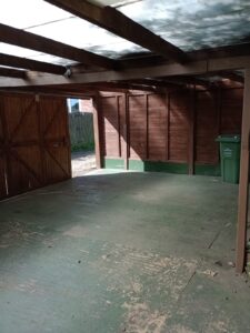Property for sale A mostly empty wooden garage with green flooring, sunlight streaming in from the open doors, and a green recycling bin against the right wall. The roof is partially translucent, allowing natural light inside. Presented by Palace Auctions