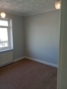 Property for sale A small, empty room with pale grey walls, a brown carpet, two hanging light fittings, a window with a radiator below it, and white trim along the skirting boards and ceiling. Presented by Palace Auctions