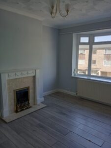Property for sale A bright, empty room with light wooden flooring, a white decorative fireplace, pale blue walls, a radiator under a large window, and a ceiling-mounted light fitting. The window shows a view of nearby buildings. Presented by Palace Auctions