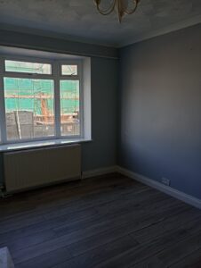 Property for sale An empty room with light grey walls, wooden floor, a large window with a radiator beneath, and a ceiling light fitting. Scaffolding and green construction netting are visible outside the window. Presented by Palace Auctions