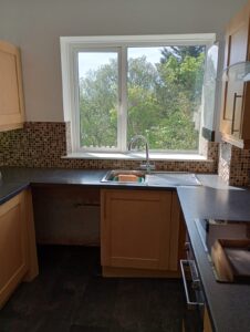 Property for sale A kitchen with wooden cupboards, black worktops, a stainless steel sink under a large window, and mosaic tile splashback. Sunlight comes through the window, showing green trees outside. Presented by Palace Auctions
