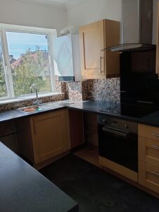 Property for sale Modern kitchen with wooden cupboards, black worktops, a built-in oven, mosaic tile splashback, and a large window above the sink letting in natural light. Trees and rooftops are visible outside. Presented by Palace Auctions