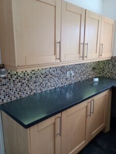 Property for sale Light wood kitchen cupboards with silver handles mounted above and below a black worktop. The splashback features small square mosaic tiles in shades of brown, beige, and grey. A power socket is visible. Presented by Palace Auctions