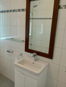 Property for sale A small white bathroom basin with a tap, set below a rectangular mirror framed in wood. The wall is tiled white with a grey mosaic accent. There is a glass shelf and a towel rail to the left of the mirror. Presented by Palace Auctions